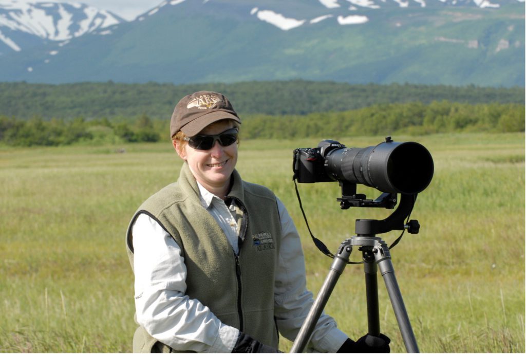 A woman with sunglasses and a hat smiles while adjusting a camera on a tripod