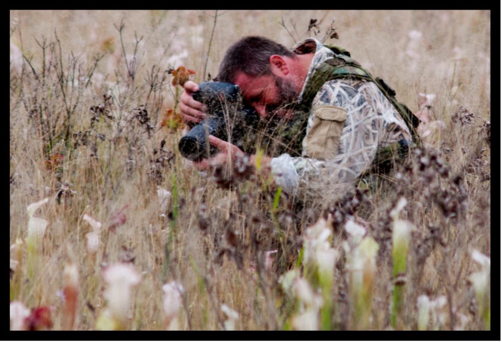 A man crouches down in thick brush while pointing a camera down at an angle